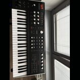Černý synthesizér s klaviaturaí, na jehož horním panelu je bílý nápis HYDRASYNTH a logo MOOG. Přístroj obsahuje množství ovládacích knoflíků, posuvní…