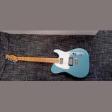 Modrá elektrická kytara s bílým pickguardem a světlou hnedou hmatníkovou deskou. Na hlavě je viditelné logo Fender. Kytara má dva snímače a kovový mo…