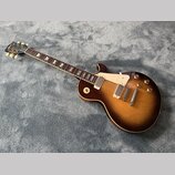 Elektrická kytara s tónem sunburst v hnědých odstínech a bílým pickguardem. Na hlavě je viditelné logo Gibson. Hриф má tmavý hmatník s trapezoidovými…