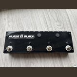 Černý obdélníkový pedalboard s nápisem ULASA & BLACK v bílém stylizovaném písmu. Na přední straně jsou čtyři kovové spínací tlačítka s bílými podložk…