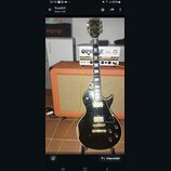 Černá elektrická kytara s tvarem singlecut a bílým rámečkem kolem těla. Na hlavě je viditelné zlaté logo Gibson. Kytara má dva humbuckery, čtyři ovlá…