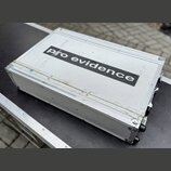 Stříbrný transportní kufr z hliníku s texturovaným povrchem. Uprostřed je černý nápis pro evidence. Povrch vykazuje známky opotřebení v podobě škrába…