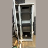 Bílá kovová serverová skříň s otevřenými dveřmi. V interiéru jsou umístěny tři černé police na různých úrovních. Povrch skříně vykazuje běžné známky…