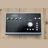 Černý audio rozhraní s kovovým šedým rámem a nápisem audient v horní části. Na předním panelu je označení iD44, čtyři otočné knofíky GAIN, velký cent…