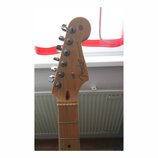 Fotografie zachycuje hlavu elektrické kytary v přírodním dřevě s chromovanými ladičkami. Na hlavě je černý nápis Fender, STRATOCASTER® a menší text C…