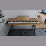 Digitální piano v dřevěném provedení s bílými a černými klávesami. Na středové liště pod klaviaturou je nápis HENRY'S. Nástroj stojí na čtyřech šedýc…