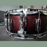 Detailní záběr na snare drum s tmavě hnědým dřevěným korpusem a chromovanými obručmi. Na chromovaném mechanismu napnutí je vyryto logo "Gretsch". Nás…