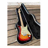 Elektrická kytara v barvě sunburst s bílým pickguardem, umístěná v černém tvrdém kufru. Na hlavě kytary je viditelné logo Fender. Nástroj má tři sním…