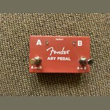 Červený kovový kytarový pedál s nápisem Fender ABY PEDAL. Na horní straně jsou bílá písmena A a B, nápis IN/OUT a dva kovové spínače s popisky A and…