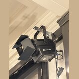 Černý světelný reflektor s nastavitelnými klapkami, který je připevněn na kovový držák. Na boku přístroje je bílý nápis ADJ ENCORE FRISO2 a symbol CE…