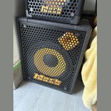 Černý basový reproduktor s kovovou mřížkou, na které je žlutý kruh a čtverec. V dolní části mřížky je umístěno žluté logo Mark bass. Nad ním je vidět…
