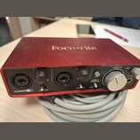 Červené audio rozhraní značky Focusrite, model Scarlett 2i2. Přední panel obsahuje dva kombinované vstupy, dva ovladače zisku s nápisy LINE INST a je…