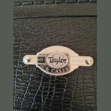 Detail kovového štítku na černém kufru s texturou kůže. Štítek obsahuje logo Taylor a nápisy QUALITY GUITARS & CASES. Je připevněn dvěma šrouby, povr…