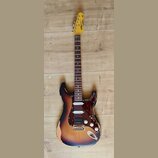 Elektrická kytara s tłem v barvě sunburst a hnědým pickguardem. Na hlavě je viditelné logo "Vanguard" a nápis "ICON Series". Tělo kytary vykazuje výr…