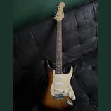 Elektrická kytara s tónem těla v přechodové barvě od žluté po tmavě hnědou a bílým pickguardem. Na hlavě hředu je viditelné logo Squier. Nástroj má t…