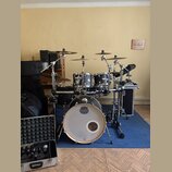 Černá bicí souprava s bílým předním zrcadlem basového bubnu, na kterém je logo MAPEX. Na stojanu je viditelný nápis Gibraltar. Souprava obsahuje něko…