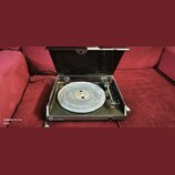 Gramofon s černým šasi a průhledným plastovým krytem, který je otevřený. Na talíři je položena vinylová deska. V levém dolním rohu šasi je bílý štíte…