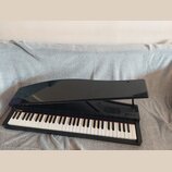 Černý digitální piano v provedení křídla s lesklým povrchem. Na levé straně nad klávesami je vidět žlutý nápis "KORG". Nástroj má standardní bílé a č…