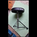 Černý bubeníkářský židle s kovovým chromovaným stativem a trojnožkou. Na sedáku je bílý nápis TAMA a pod ním text THE STRONGEST NAME IN DRUMS. Povrch…