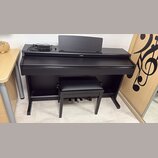 Černý digitální piano v černém nábytkovém stojanu s přívodním kabelem a doprovodnou černou stolečkou. Na přední straně je umístěno logo YAMAHA. Nástr…
