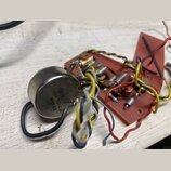 Fotografie zobrazuje elektronickou součástku v kovovém pouzdře připojenou k červené desce s dalšími komponenty a barevným zapojováním kabelů. Na kovo…
