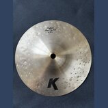Kulatý činely z kovu s viditelným broušením povrchu. Na horní části je nápis „Custom Dark Splash 8"/20 cm“ a v dolní části velké černé logo „K“. V pr…