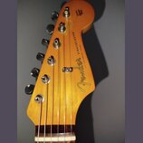 Fotografie zachycuje hlavu elektrické kytary z přírodního dřeva s viditnou sletou. Na povrchu je černý nápis Fender a STRATOCASTER, doplněný menšími…