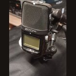 Černý digitální záznamový přístroj značky ZOOM s kovovou mřížkou mikrofonů v horní části. Zařízení má malý LCD displej a je připevněno na černý stati…