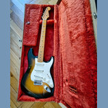 Elektrická kytara s tónem sunburst a bílým pickguardem v otevřeném kufru s červenou plišovou výstelkou. Na hlavě hředu je viditelné logo Fender. Kyta…