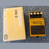 Žlutý kytarový efekt BOSS OS-2 Over Drive/Distortion v originální žluté krabici. Na pedálu jsou čtyři ovládací knoflíky, červená kontrolka CHECK a ná…