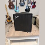 Černý kytarový kombo zesilovač s logem Fender v horní levé části mřížky. Zesilovač má hranatý tvar, černé povrchové provedení a nahoře integrovanou r…