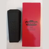 Černý wah-wah pedál s texturou na povrchu a vyraženým logem VOX. Vedle něj je červená papírová krabice s logem VOX AMPLIFICATION LTD. a nápisy V845,…