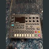Digitální syntetizátor Elektron Digitone v šedé barvě. Na horní části je nápis Polyphonic Digital Synthesizer a logo Elektron. Zařízení obsahuje disp…