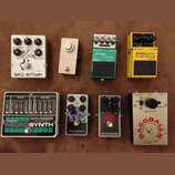 Fotografie zobrazuje osm kytarových efektů rozložených ve dvou řadách. V horní řadě jsou pedály s nápisy BASS AUTOWAH, DFZ, Boss Bass Comp BC-1X a Bo…