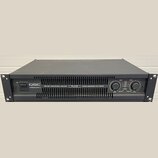 Černý rackový zesilovač s nápisy QSC POWERLIGHT 2, 1800 WATT PROFESSIONAL AMPLIFIER a PL218 CUSTOM ENGINEERED FOR TOURING. Na pravé straně jsou dva o…