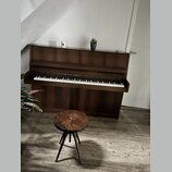 Hnědé dřevěné piano s klasickou klaviaturou. Před nástrojem stojí malá trojnožka dřevěná stolička s kulatým sedákem. Na horní desce piana jsou umístě…