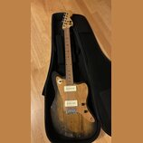 Elektrická kytara s přírodním dřevěným povrchem těla a hnedým hmatníkem, umístěná v černém obalu. Na hlavě je viditelné logo "Fender". Kytara má dva…