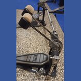 Fotografie zobrazuje bicí pedál s černým kovovým základem a šedým stupněm. Na stupni je viditelné logo Pearl. Pedál má dvě nastavitelé beatery s bílý…