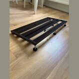 Černý kovový pedalboard s mřížkovým povrchem z gumových nebo plstěných pásů. Na boční hraně je bílý nápis „Harley Benton“ s logem. Nástroj stojí na č…