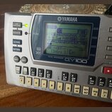 Stříbrný sekvenční modul s displejem a klávesovou ovládací. Na horní straně je logo YAMAHA a pod displejem nápis MUSIC SEQUENCER QY100. V levé části…