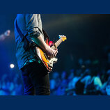 Fotografie zachicuje detail elektrické kytary v rukou hudebníka. Nástroj má tělo s barevným přechodem (sunburst), bílý pickguard a světle hnědý hриф…