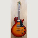 Elektrická kytara s tłem v barvě sunburst a zlatým hardwarem. Na hlavě je nápis johnson. Kytara má dva humbuckery, černý plechtrum a čtyři černé ovlá…