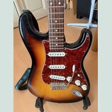 Elektrická kytara s tělem v barvě sunburst a hnědým hmatníkem. Nástroj má červený vzorovaný pickguard a tři bílé snímače. Na těle nejsou viditelné žá…