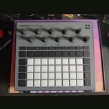 Šedý sekvenční sampler s názvem CIRCUIT RHYTHM a logem Novation v pravém horním rohu. Zařízení obsahuje několik otočných knoflíků, řadu malých tlačít…