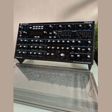 Černý syntetizátor s nápisy novation a PEAK. Na spodním okraku je text OXFORD-OSCILLATORS a EIGHT-VOICE POLYPHONIC SYNTHESISER. Přístroj obsahuje num…