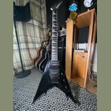 Černá elektrická kytara s tělem ve tvaru V. Na hlavě je viditelné logo Jackson. Kytara má dva humbuckery, fixační most a hриф s vkládanými značkami.…