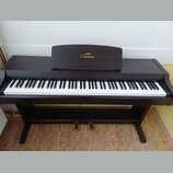 Digitální piano v tmavě hnědé barvě s dřevěnou strukturou. Na notovém pultu je zlatý nápis YAMAHA a Clavinova. Nástroj má standardní klávesnici s bíl…
