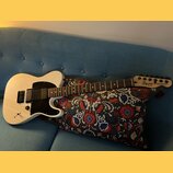 Bílá elektrická kytara s černým pickguardem a hnedSEDým hmatníkem. Na hlavě je vidět logo Squier Fender a nápis TELECASTER®. Nástroj leží na vzorovan…