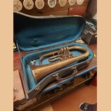 V otevřeném kufru s modrým vnitřkem leží mosazný žiletový nástroj (flugelhorn). Povrch kovu je matný s viditelnými oxidacemi a známky opotřebení. Na…