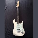 Bílá elektrická kytara typu Stratocaster s jasně zeleným pickguardem. Na hlavě hředu je viditelné logo Fender. Nástroj vykazuje známky používání, jso…
